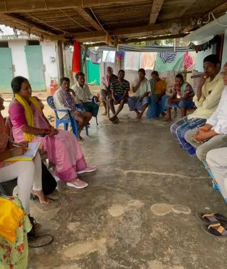 A village meeting in a courtyard: women organisers seated on one side taking notes, village elders on the other.