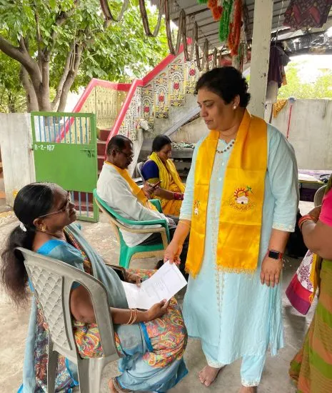 Krishna Priya handing a letter to a seated woman during a household meeting.
